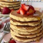 Vegan Bisquick pancakes stacked on a pink plate with chopped strawberries piled on top and maple syrup being poured. More pancakes and strawberries are in the background. Text reads "Vegan" in fancy cursive lettering and "BISQUICK PANCAKES" in bold block lettering.