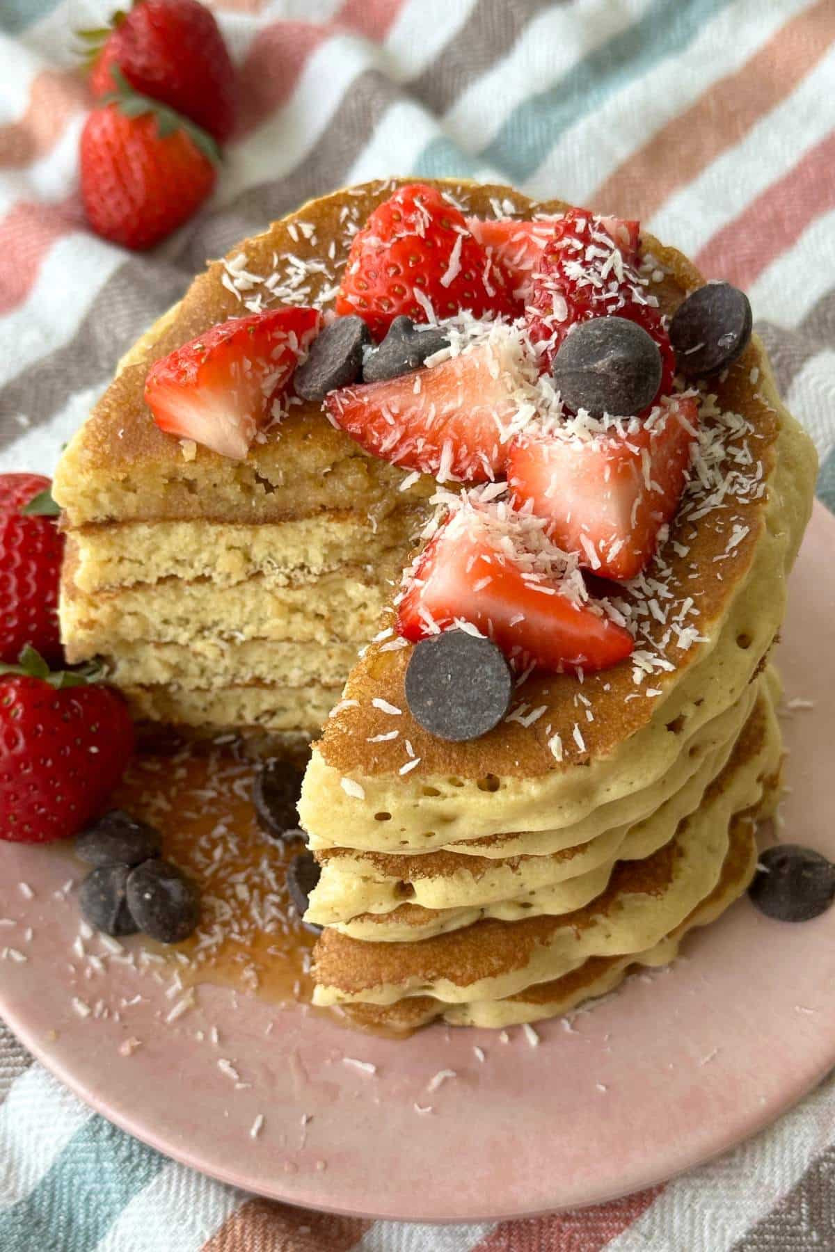 Stack of pancakes with strawberries, chocolate chips, and coconut shreds on top. A piece is cut out of the stack and maple syrup is pooling on the plate.