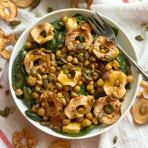 Vegan fall harvest salad in a bowl on top of a white cloth with apple chips and pepitas scattered around.