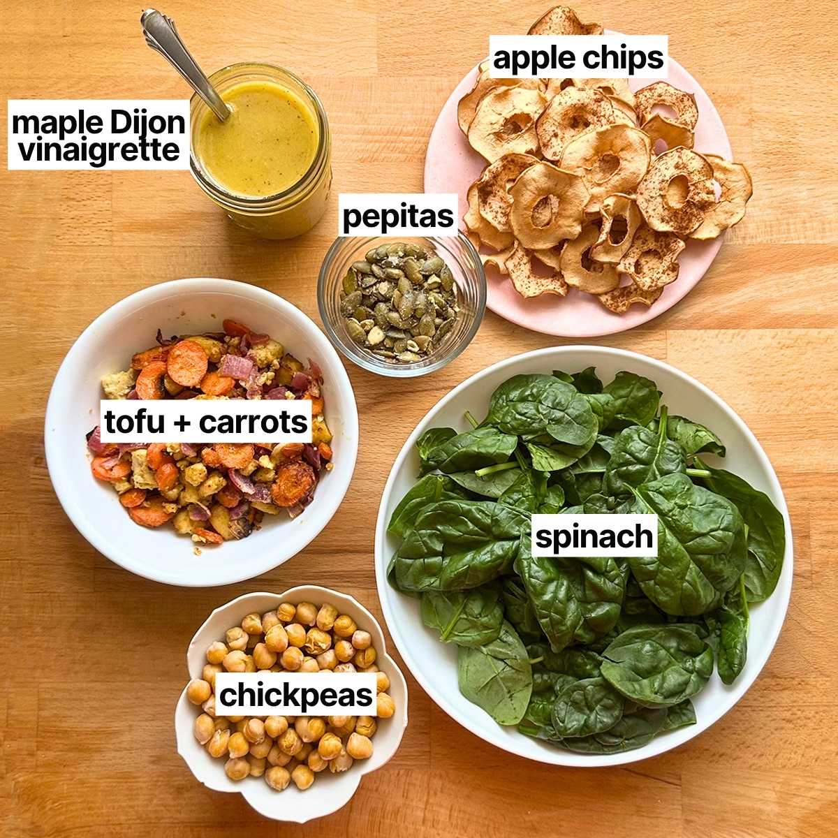 Ingredients to make vegan fall harvest salad with corresponding labels: maple dijon vinaigrette, pepitas, apple chips, roasted tofu and carrots, spinach, and chickpeas.