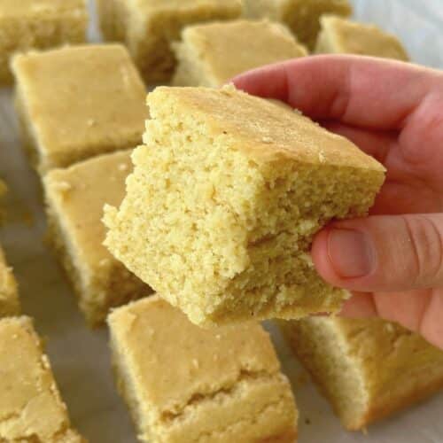 Cube of vegan cornbread being held by a hand with more cornbread in the background.