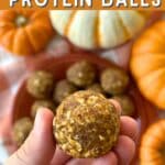 A hand holding 1 pumpkin protein ball. In the background there is a plate of the balls with 4 pumpkins surrounding it. Text reads "Vegan" in fancy lettering and "PUMPKIN PROTEIN BALLS" in bold block lettering.