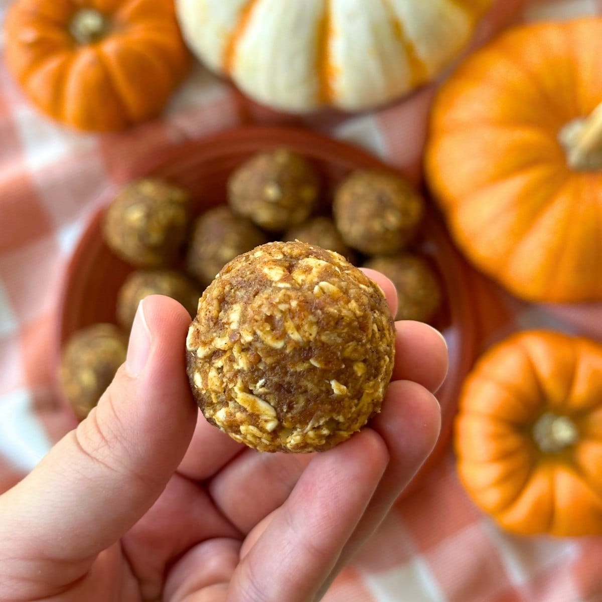 A hand holding 1 pumpkin protein ball. In the background there is a plate of the balls with 4 pumpkins surrounding it.