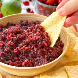 Tortilla chip dipped into cranberry jalapeno salsa with chips and cranberries in the background.