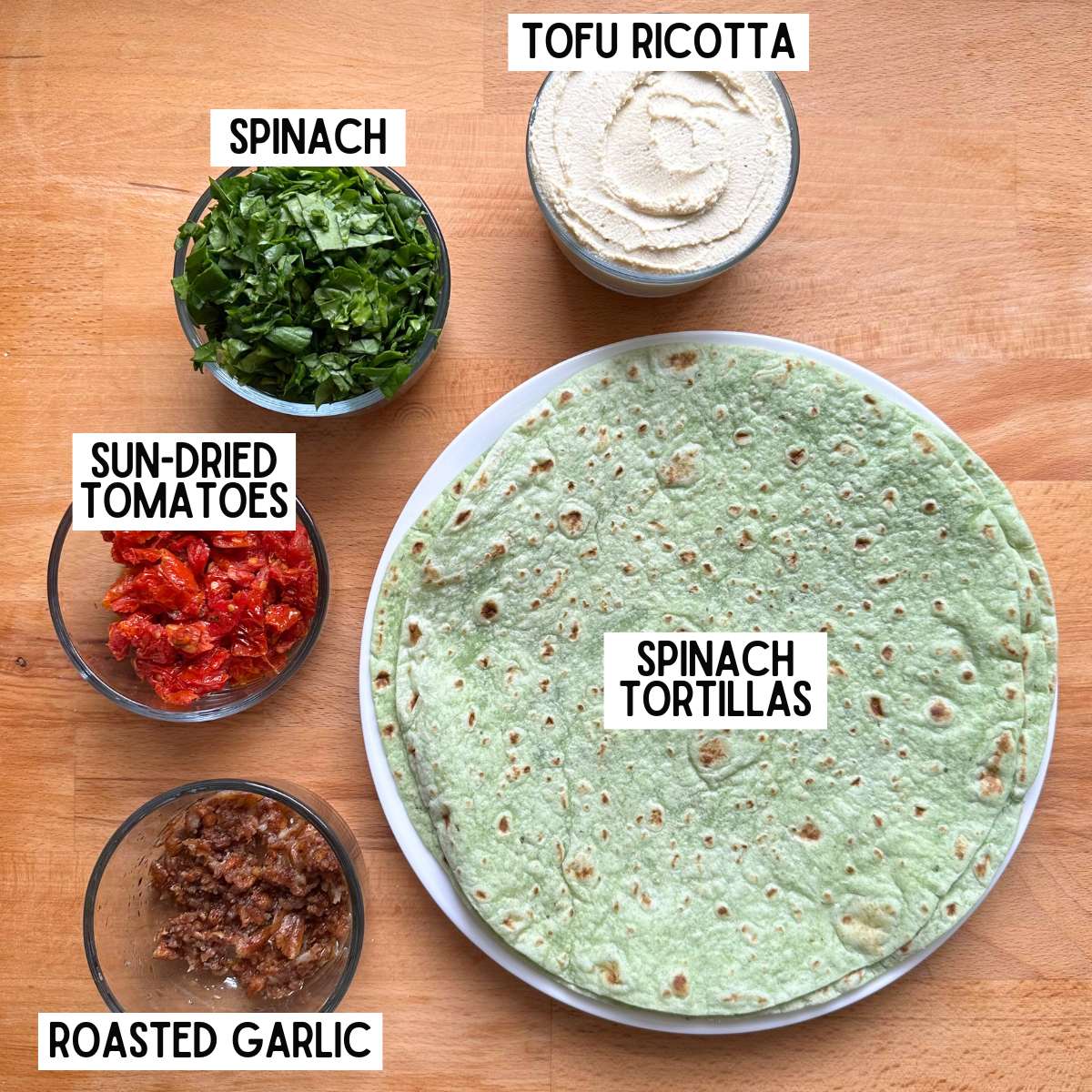 Ingredients to make vegan pinwheels with corresponding labels: tofu ricotta, spinach, sun-dried tomatoes, roasted garlic, and spinach tortillas.
