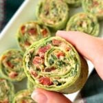 Vegan pinwheel with green tortilla and tofu, sun-dried tomato filling on a plate with hand holding up one piece.