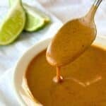Spoon with almond sauce dripping off of it into a small bowl with almond sauce. Lime wedges are in the background on top of a white cloth napkin.