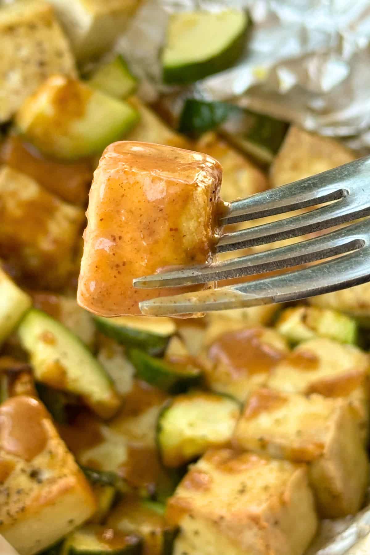 Piece of tofu on a fork dipped in almond sauce with more tofu and zucchini in the background drizzled with the sauce.