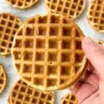 Mini Bisquick waffle held in a hand with more vegan waffles in the background on a white background.