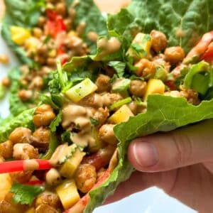 Chickpea lettuce wrap being held, filled with chickpeas, red pepper, mango, cilantro, green onion and drizzled with peanut sauce.