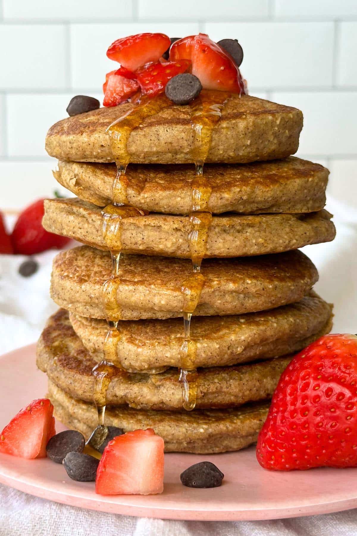 Tall stack of vegan oatmeal blender pancakes on a pin plate. Strawberries and chocolate chips are piled on top. Maple syrup is running down the stack.