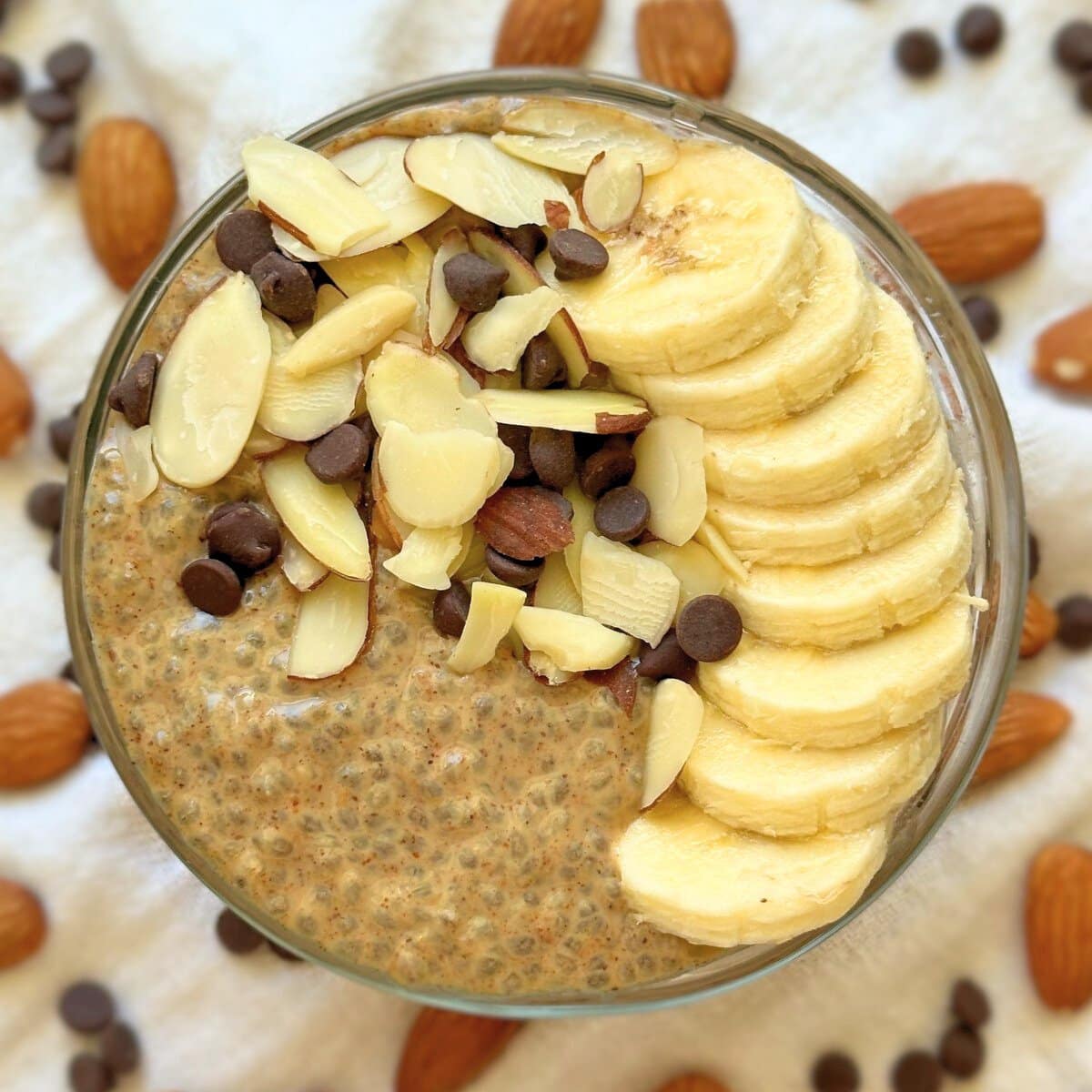 Almond butter chia pudding topped with sliced banana, almonds, and chocolate chips. More almond and chocolate chips are in the background.