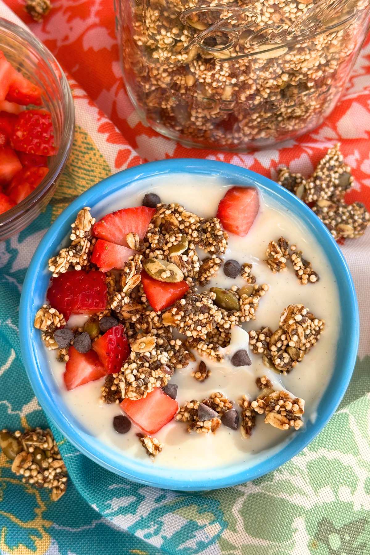 Bowl of yogurt with quinoa granola, strawberries, and chocolate chips. The background is a floral napkin, a bowl of strawberries, and a jar of granola.