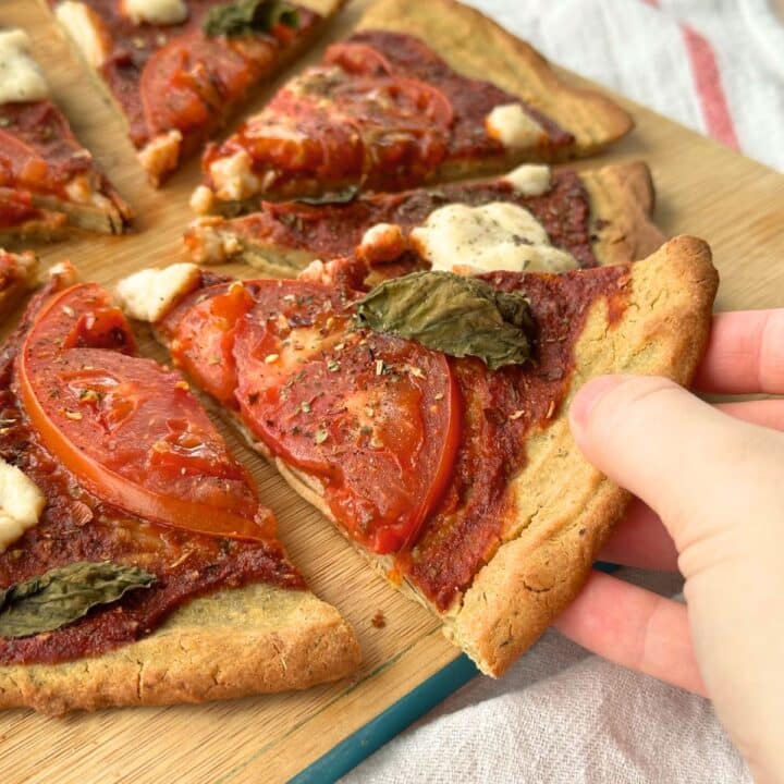 Hand taking a piece of vegan quinoa pizza with sliced tomato, basil, and vegan mozzarella from a sliced pizza pie.