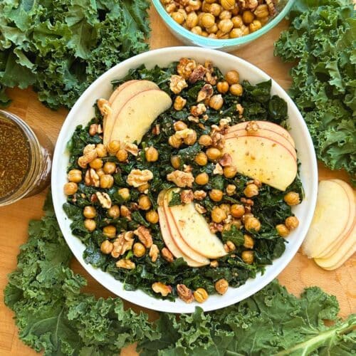 Kale salad in a bowl with apples, walnuts, and chickpea on top. Around the bowl is more kale, chickpeas, apples, and the balsamic dressing.