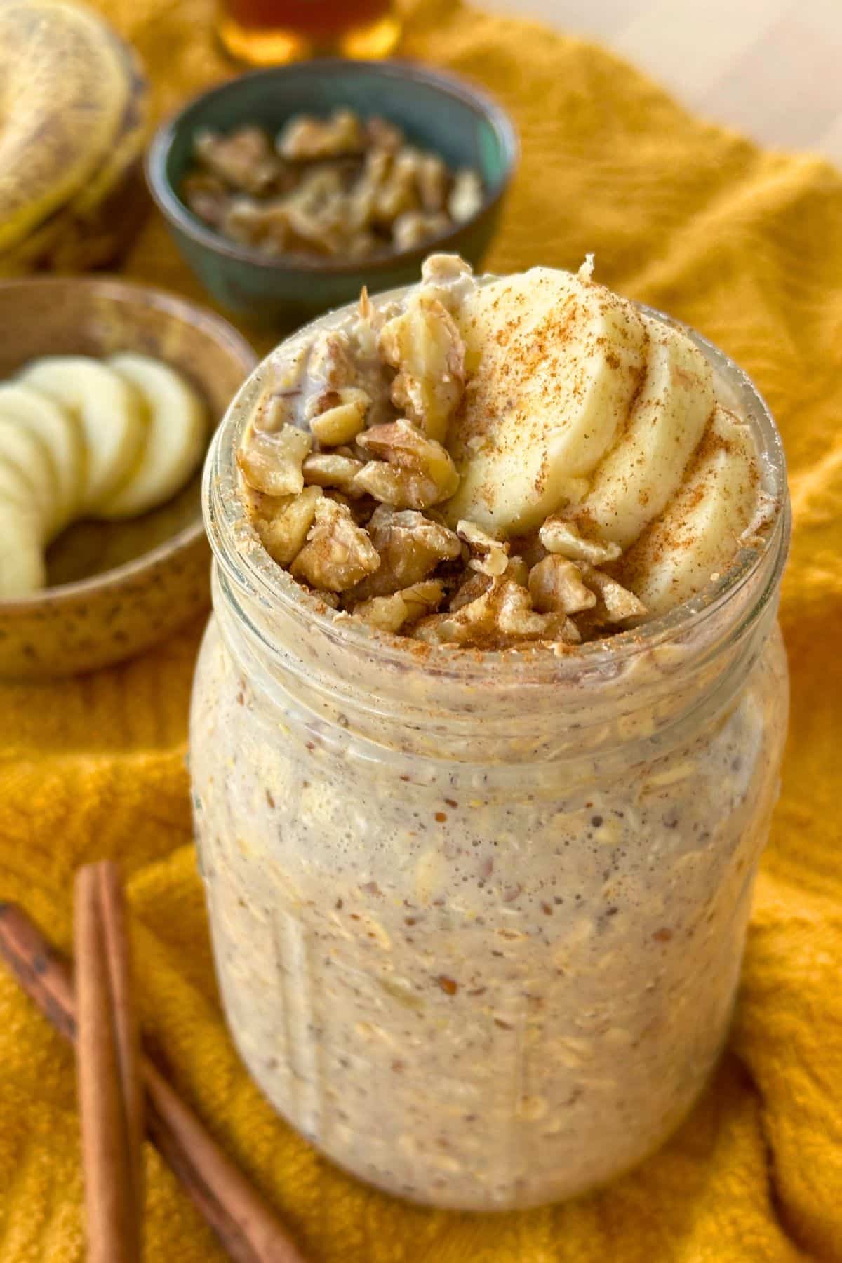 Mason jar of vegan banana bread overnight oats topped with sliced banana and walnuts; the background includes more walnuts, banana, sticks of cinnamon, and maple syrup.