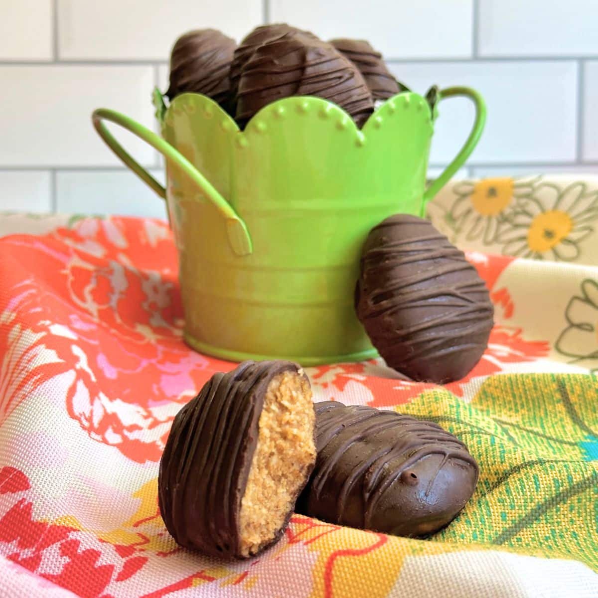 Chocolate almond butter egg with a bite taken out of it resting on top of another chocolate egg; more chocolate eggs are in a green basket in the background on top of  a floral napkin.