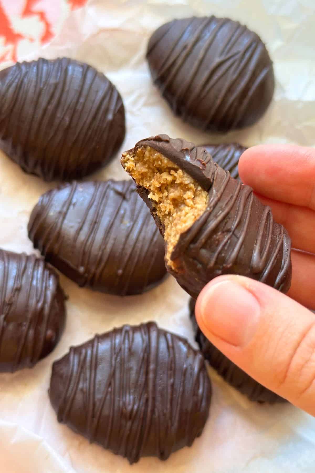 Hand holding up a piece of an almond butter egg with a bite taken out of it, more chocolate eggs are in the background.