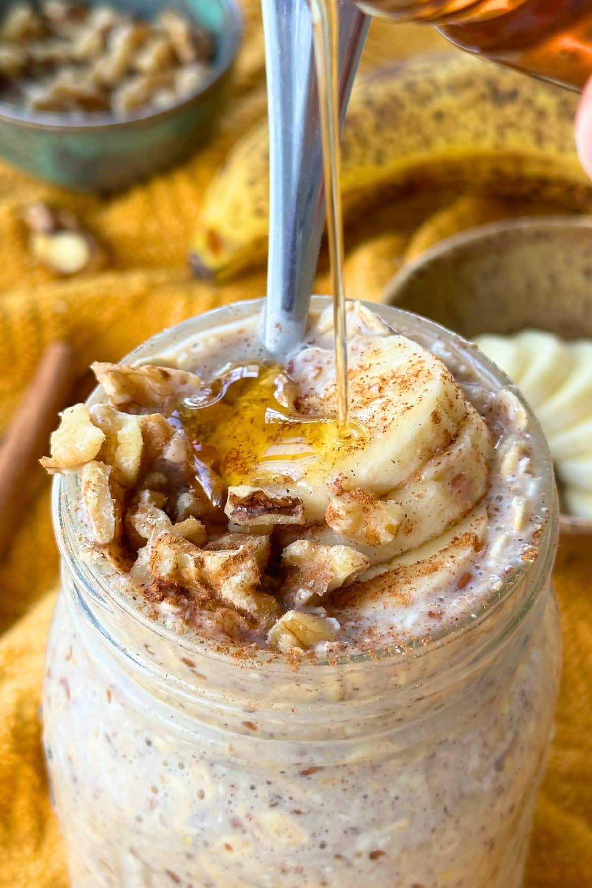 Maple syrup being poured on top of banana overnight oats topped with sliced banana, chopped walnut, and a sprinkle of cinnamon; more of the toppings are in the background on top of a mustard yellow napkin.