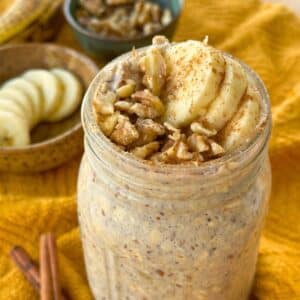 Vegan banana bread overnight oats in a glass jar topped with sliced banana, chopped walnut, and a sprinkle of cinnamon; more of the toppings are in the background on top of a mustard yellow napkin.