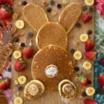 Vegan easter bunny pancake with a bunny tail, paws, and ears on a board with strawberries, chocolate chips, banana slices, and coconut flakes.