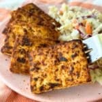 Blackened tofu pieces stacked on a plate with vegan coleslaw, a fork is holding up the first piece of tofu.