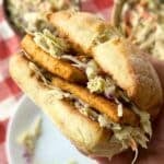 Blackened tofu and creamy vegan coleslaw sandwich being held up by a hand with more coleslaw in bowls in the background on top of a red and white gingham napkin.