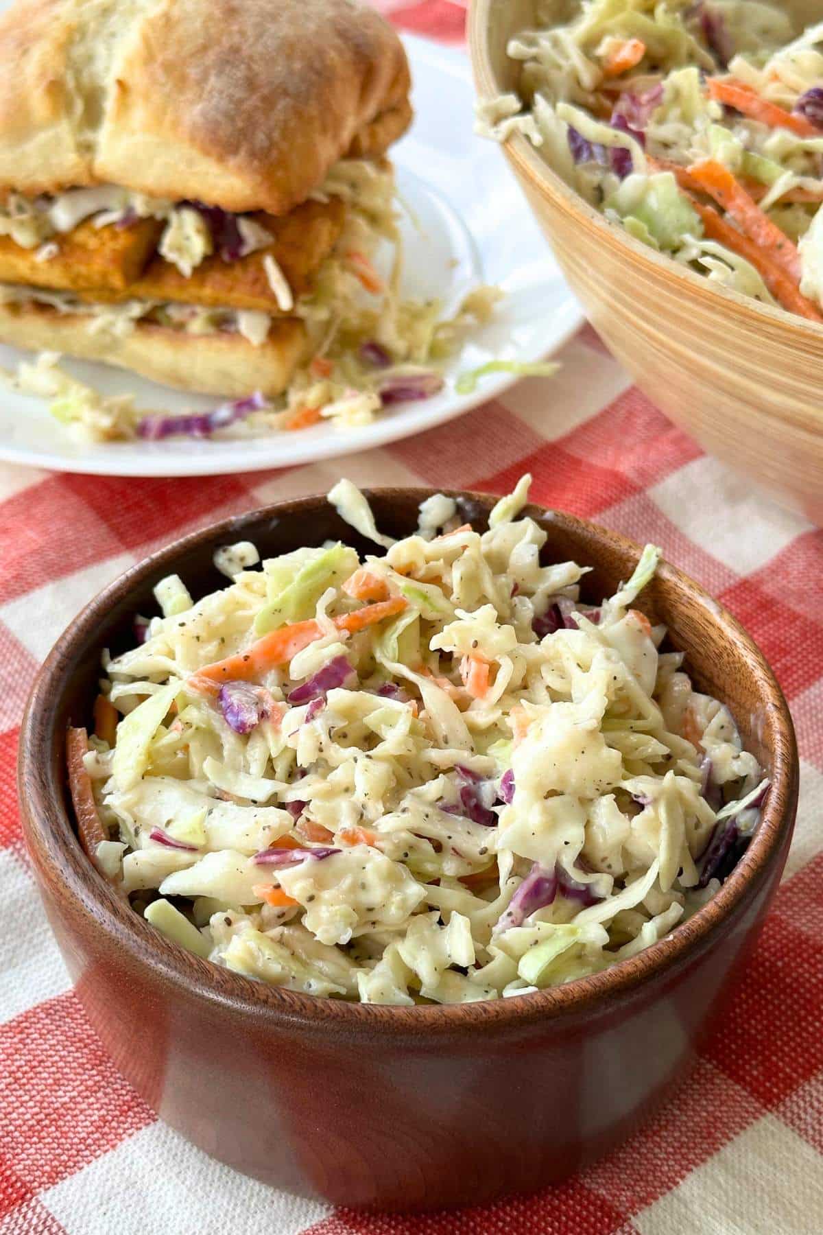 Creamy coleslaw in a small wooden bowl, a sandwich with coleslaw is in the background on top of a red and white gingham napkin.