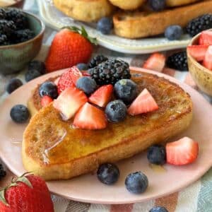 Two pieces of vegan French toast on a pink plate with berries; a stack of toast is in the background with more toppings in small bowl and scattered around.