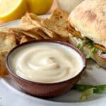 Vegan mayo in a ceramic bowl with chips and a sandwich in the background.