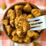 Nutritional yeast tofu nugget on a fork with more in the background in a bowl.