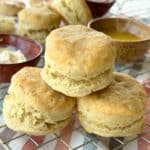 One vegan buttermilk biscuit stacked on top on of 2 other biscuits, more biscuits are scattered in the background with butter.