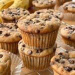 Two vegan gluten-free banana muffins with chocolate chips stacked on top of one another with more muffins and bananas in the background.