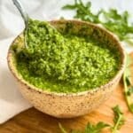 Vegan arugula pesto in a cream colored ceramic bowl with a spoon, arugula leaves are in the background.