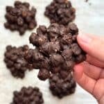 Hand holding a chocolate covered chickpea cluster with salt on top, more clusters are in the background.