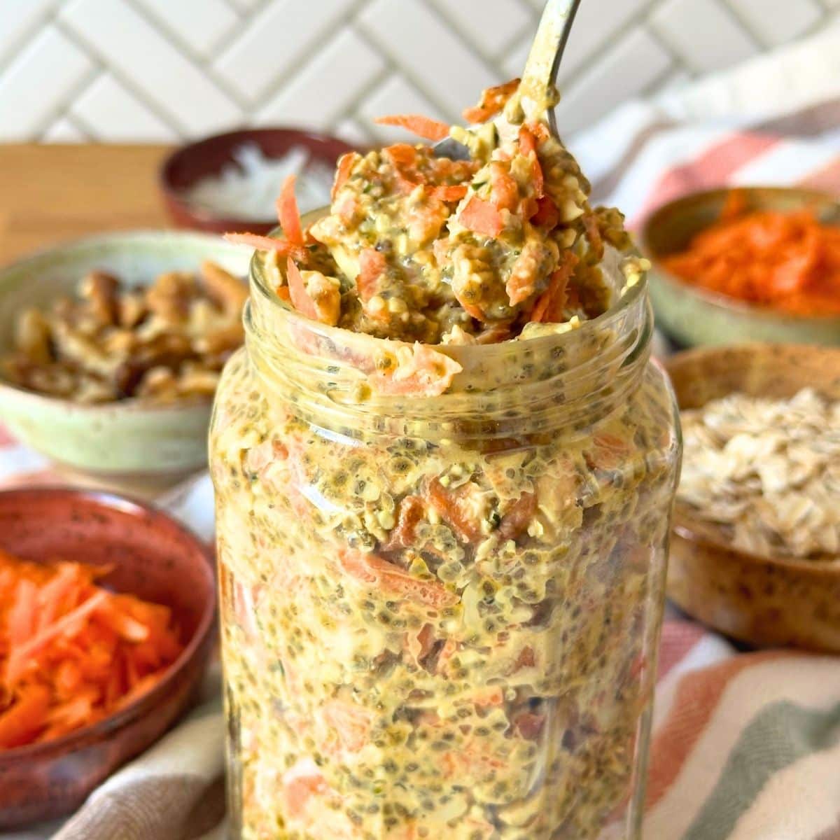 Carrot cake overnight oats in a glass jar with a spoon taking out some of the oatmeal, bowls of shredded carrot, walnuts, oats, and coconut are in the background.