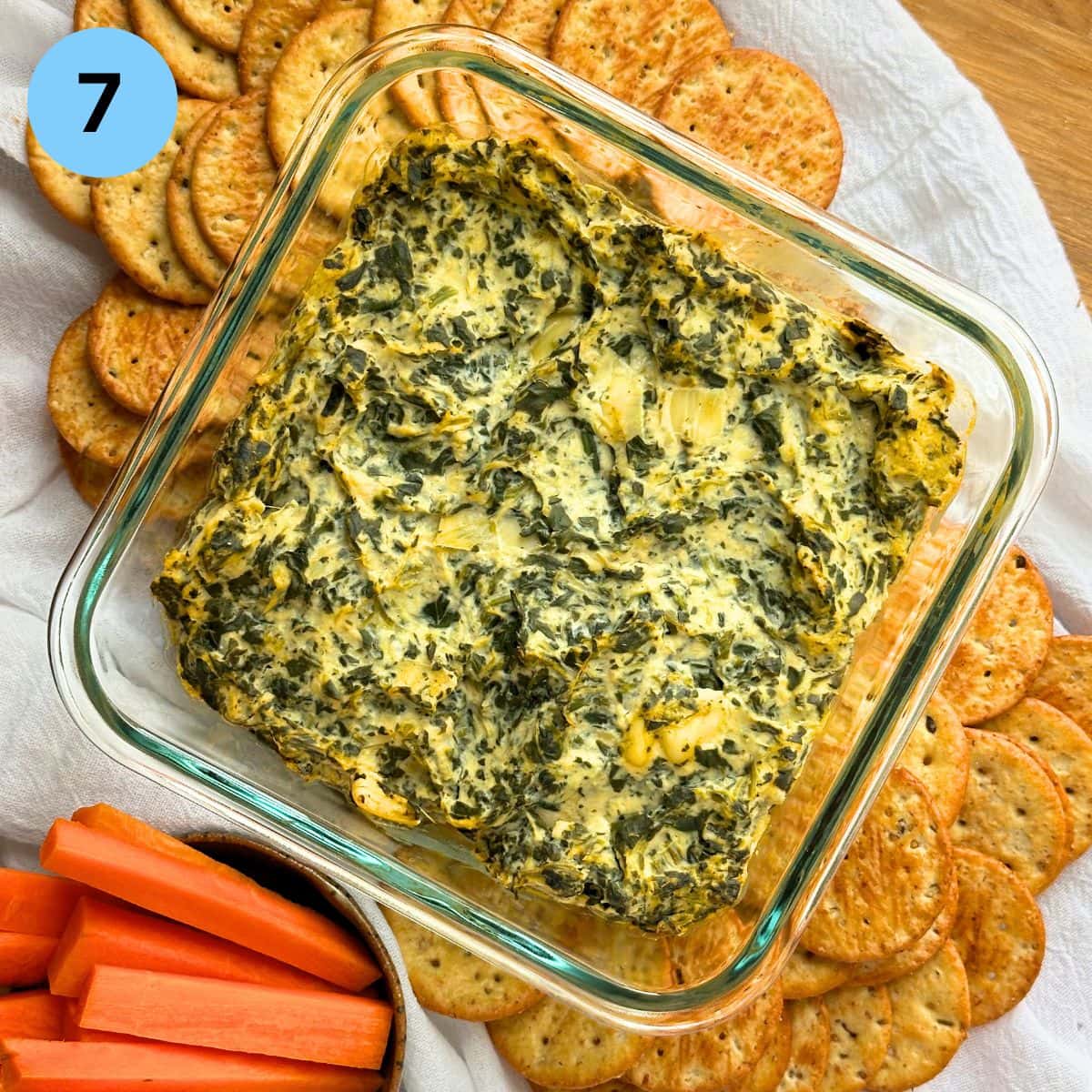Baked spinach artichoke dip surrounded by crackers and chopped carrot sticks.