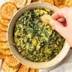 Cracker being dipped into a ceramic bowl filled with vegan spinach artichoke dip, surrounded by crackers.