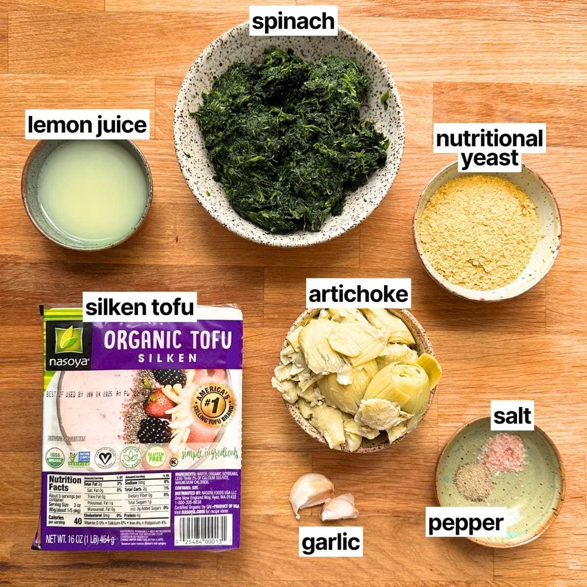 Ingredients needed to make vegan spinach and artichoke dip with labels: spinach, lemon juice, nutritional yeast, silken tofu, artichoke, garlic, salt, and pepper.