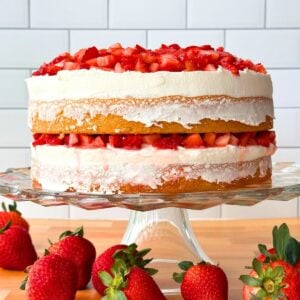 Two layer lemon cake with chopped strawbrries and lemon buttercream on a cake stand, surrounded by fresh strawberries.