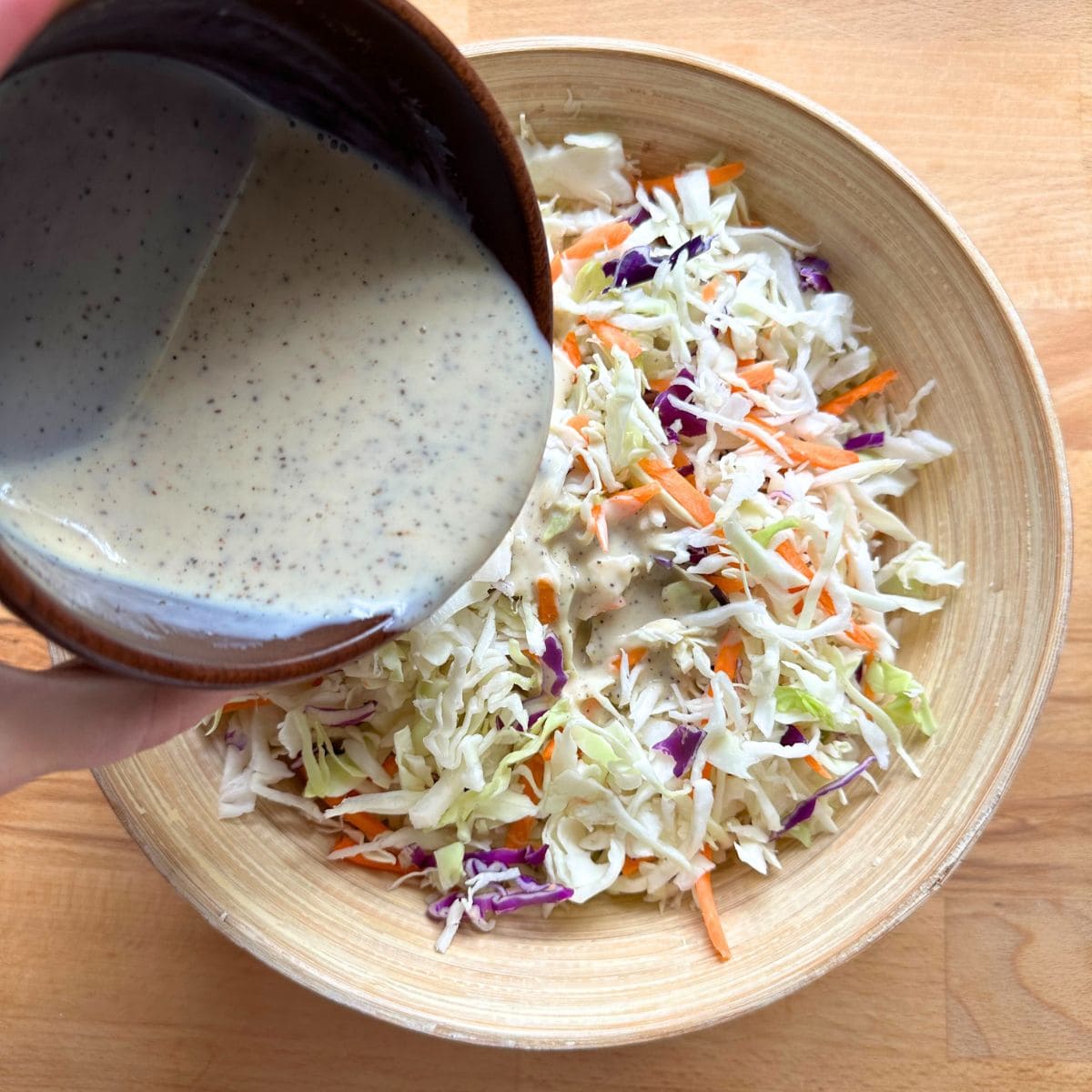 Pouring creamy silken tofu coleslaw dressing over a bowl with shredded cabbage and carrot.