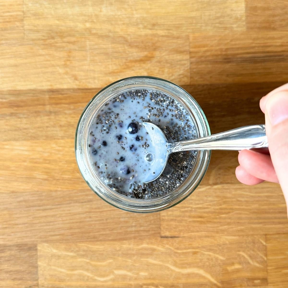 Mixing chia seeds, soy milk, and frozen blueberries in a jar with a spoon.