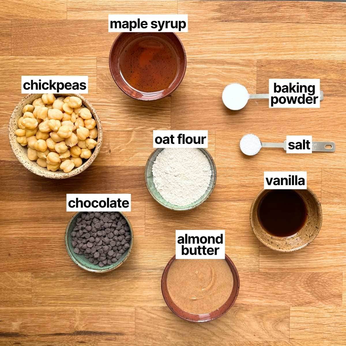 Ingredients needed to make chickpea cookies with labels: maple syrup, chickpeas, oat flour, baking powder, salt, chocolate, almond butter, and vanilla.