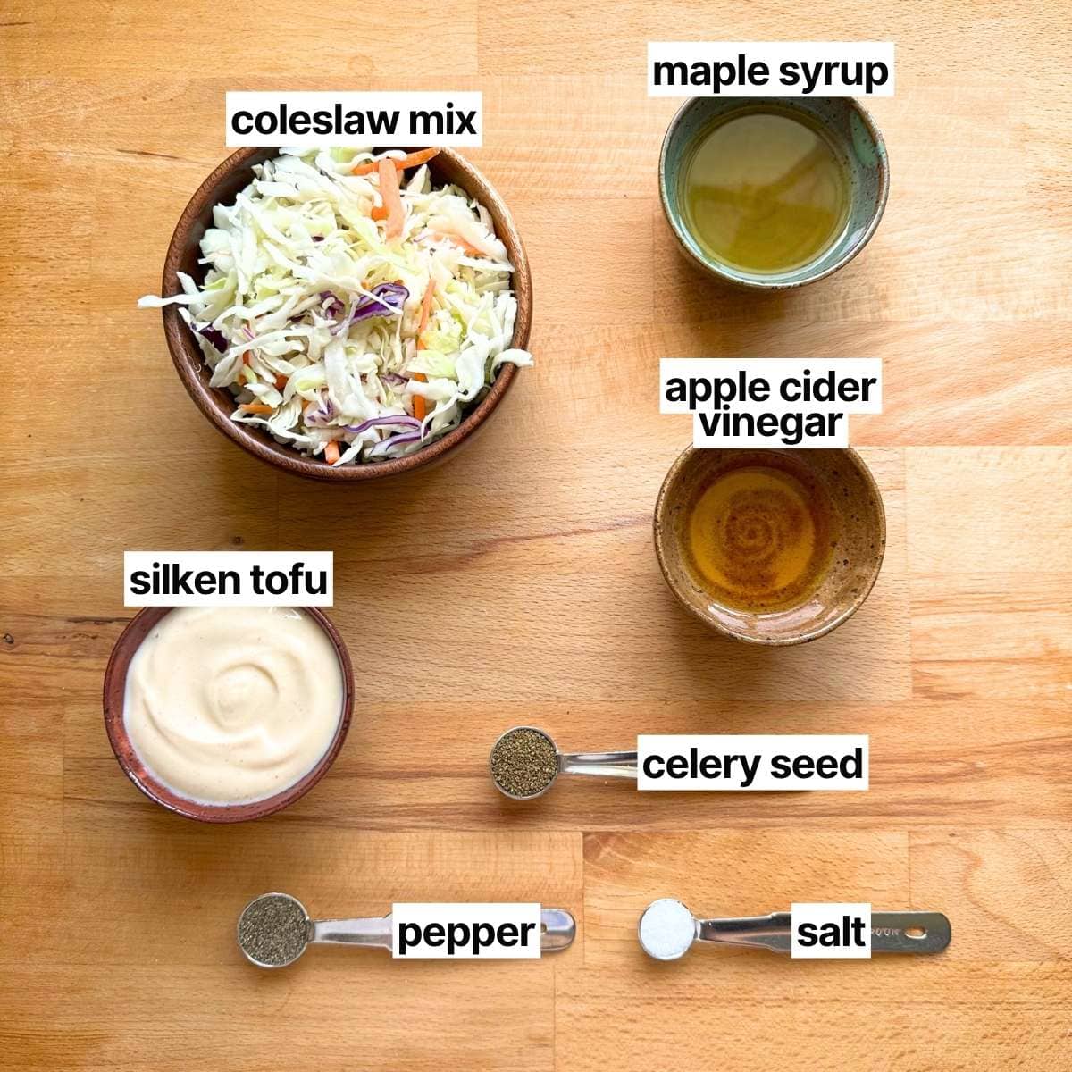 Ingredients needed to make coleslaw with labels: coleslaw mix, maple syrup, silken tofu, apple cider vinegar, celery seed, pepper, and salt.