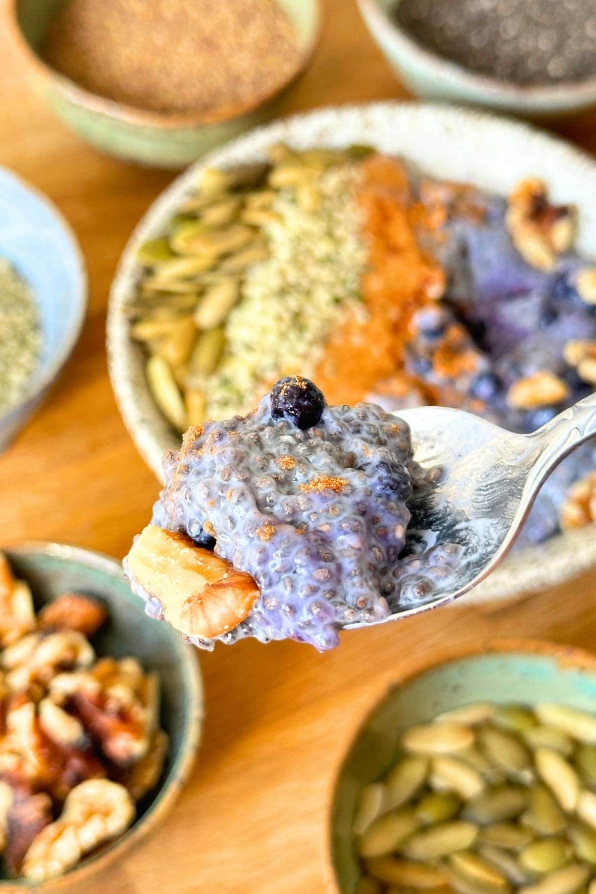 Spoonful of chia pudding with blueberry and walnut, background includes bowls of ingredients for chia pudding.