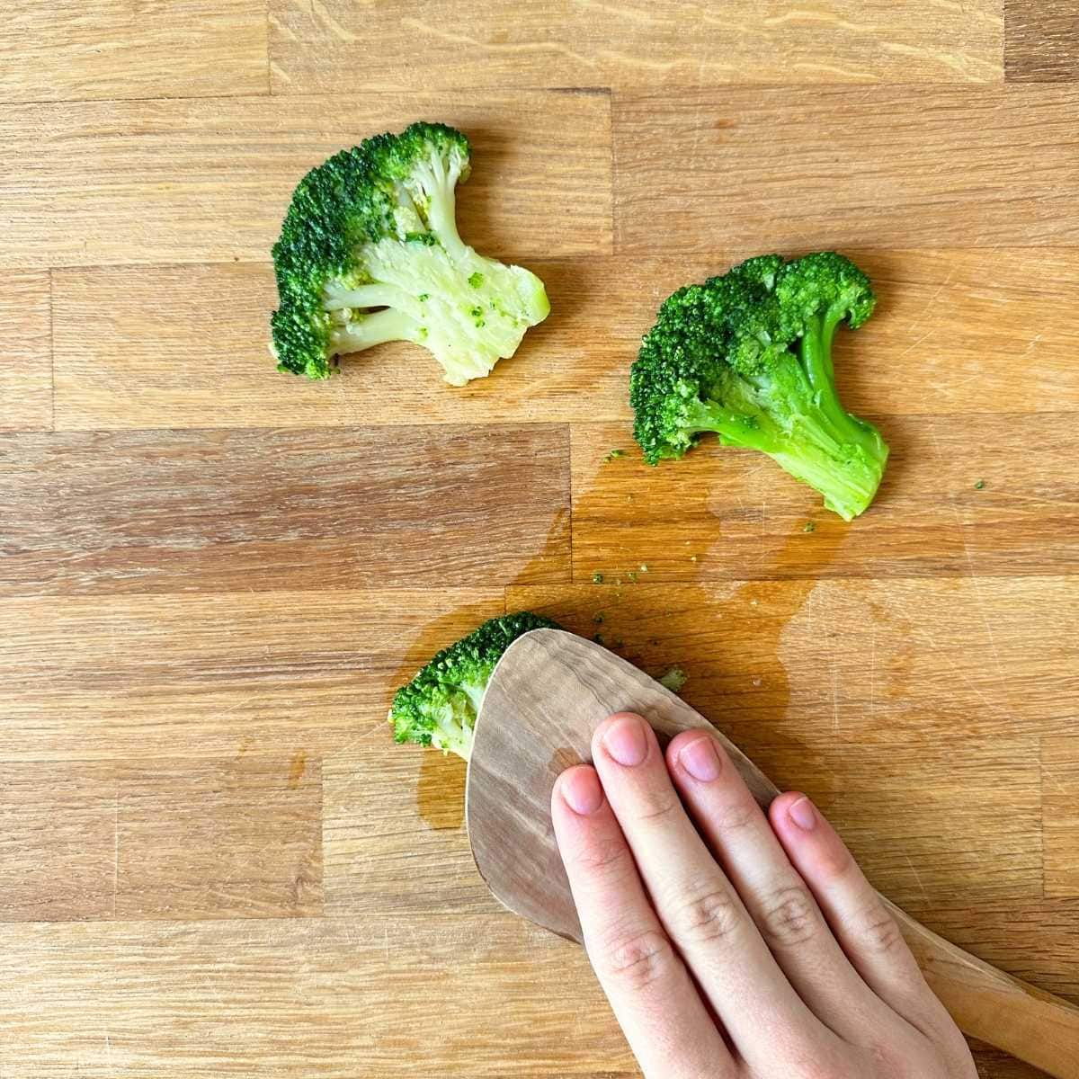 Smashing broccoli floret with a wooden spoon on a wooden cutting board.