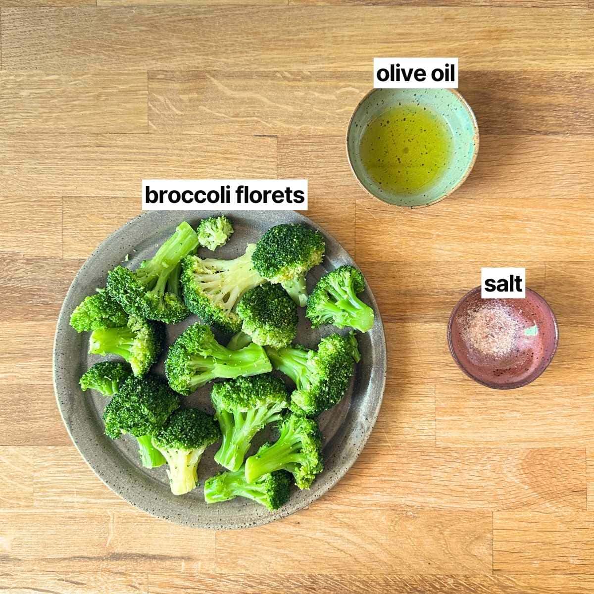 Plate of broccoli florets, bowl of olive oil, and bowl of salt on a wooden board.