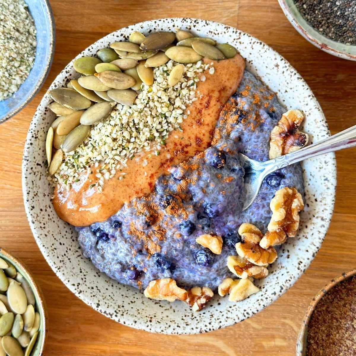 Chia pudding in a ceramic bowl topped with pumpkin seed, hemp hearts, almond butter, walnuts, and cinnamon.