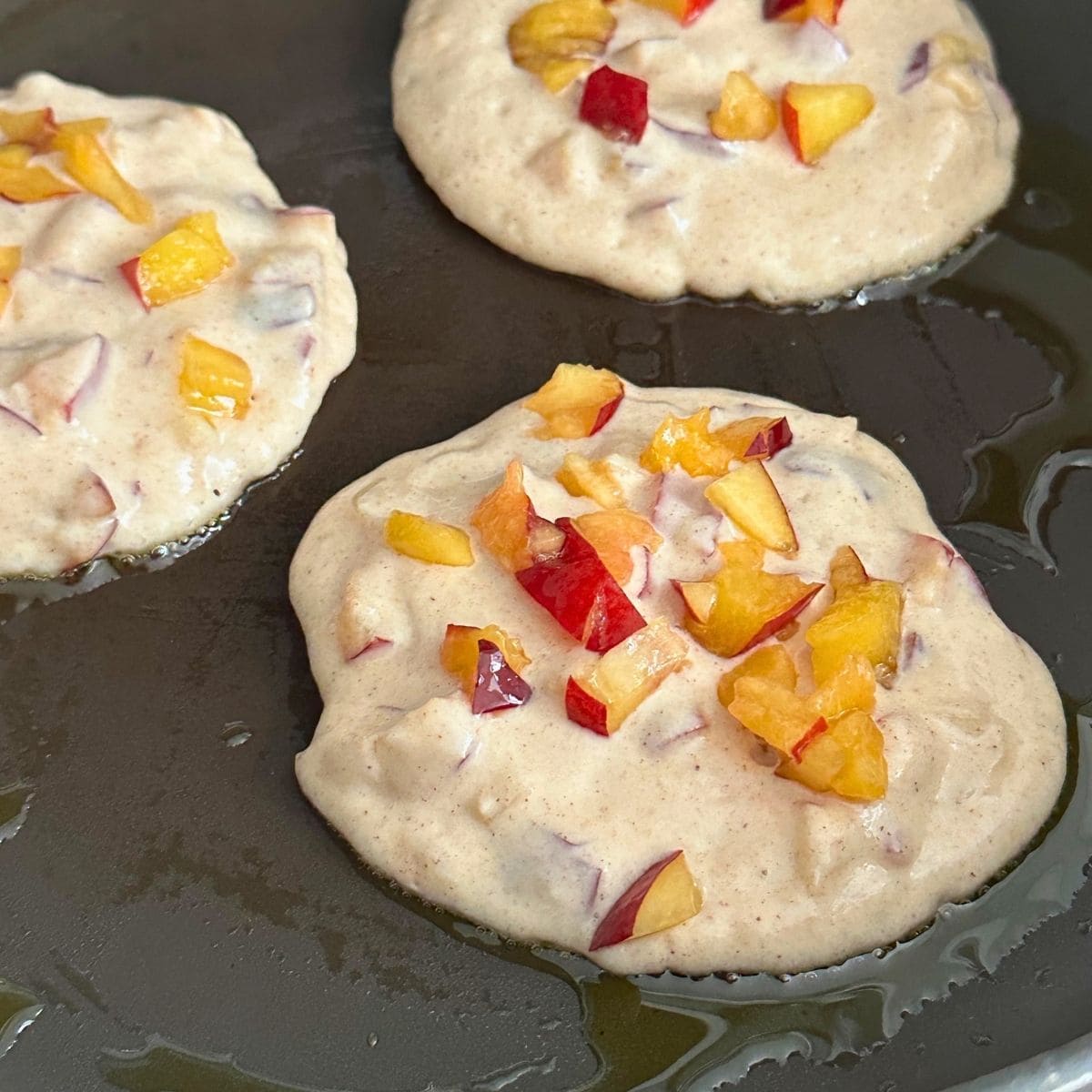 Three summer pancakes with chopped peaches on top cooking in a pan with oil.