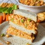 Two halves of a sandwich filled with vegan buffalo chickpea salad stacked on top of one another on a plate with more buffalo chickpeas int he background with sliced carrots, celery, and crackers.